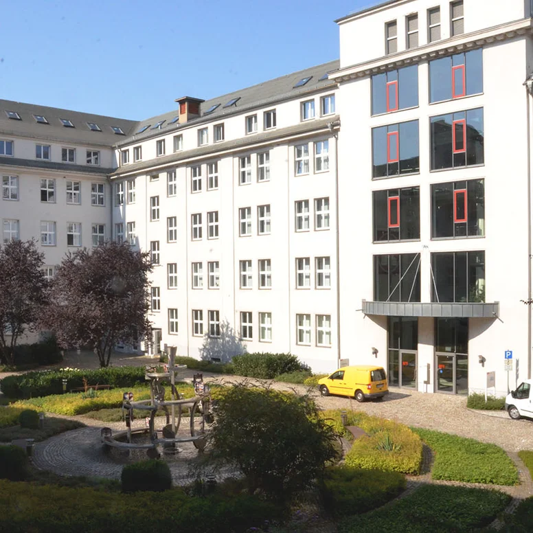Die Außenansicht des Schulgebäudes in der Kauffahrtei 25 in 09120 Chemnitz. Das Gebäude ist U-förmig gebaut und umschließt einen gepflasterten Innenhof. Im Hof befinden sich Pflanzen, Bäume und ein Springbrunnen. Ein gelbes und ein weißes Auto parken vor dem verglasten Eingangsbereich.