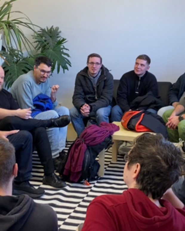 Mehrere Personen sitzen in einem Kreis in einem hellen Aufenthaltsraum und tauschen sich miteinander aus. Die Gruppe ist auf Sofas und Sesseln um einen kleinen Tisch versammelt, umgeben von zahlreichen Zimmerpflanzen.