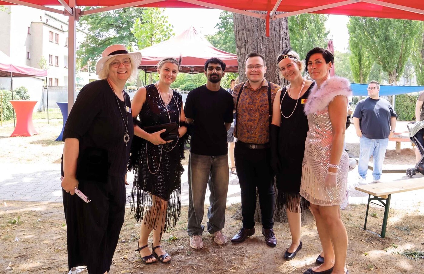 Sechs Lernbegleiter:innen und Auszubildende zur Erzieher:in posieren fröhlich unter einem roten Festzelt beim Kitafest. Sie tragen Mode der 1920er Jahre wie Paillettenkleider, Perlen, Federboas und Hosenträger.