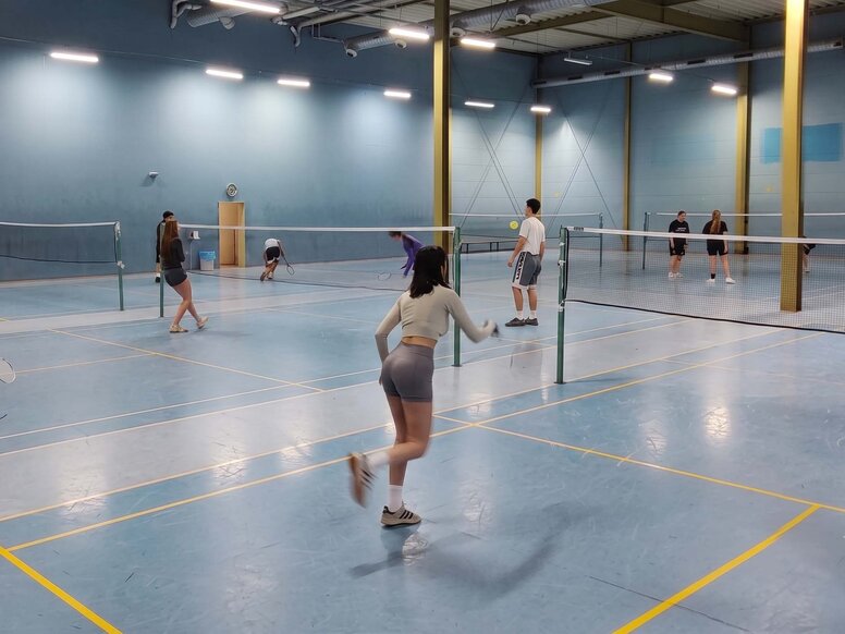 Mehrere Auszubildende zur Physiotherapeut:in spielen während des praktischen Unterrich Badminton in einer großen, gut beleuchteten Sporthalle mit blauem Bodenboden.