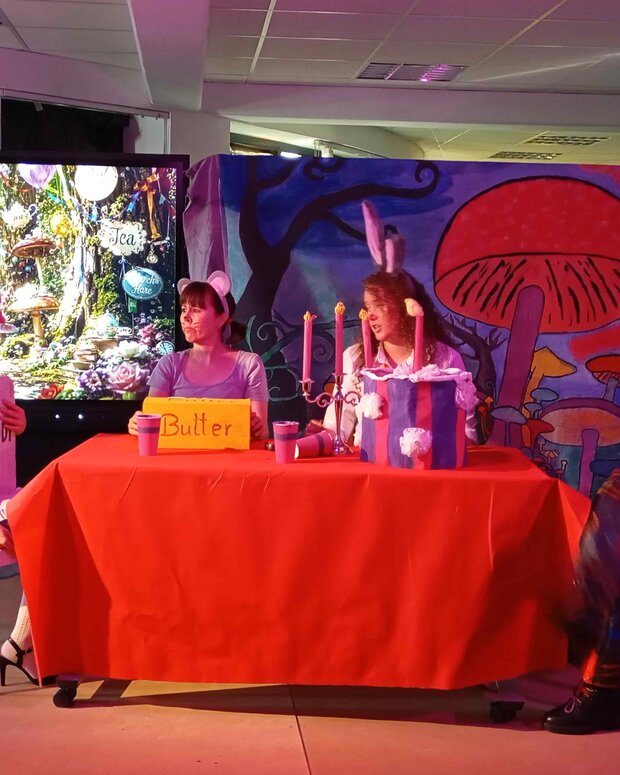 Theaterszene im Stil von „Alice im Wunderland“: Vier kostümierte Personen sitzen an einem rot gedeckten Tisch vor einer fantasievollen Kulisse mit großen Pilzen. Im Hintergrund sind ein Bildschirm mit einem märchenhaften Gartenmotiv sowie ein Schild mit der Aufschrift „Butter“ auf dem Tisch zu sehen.