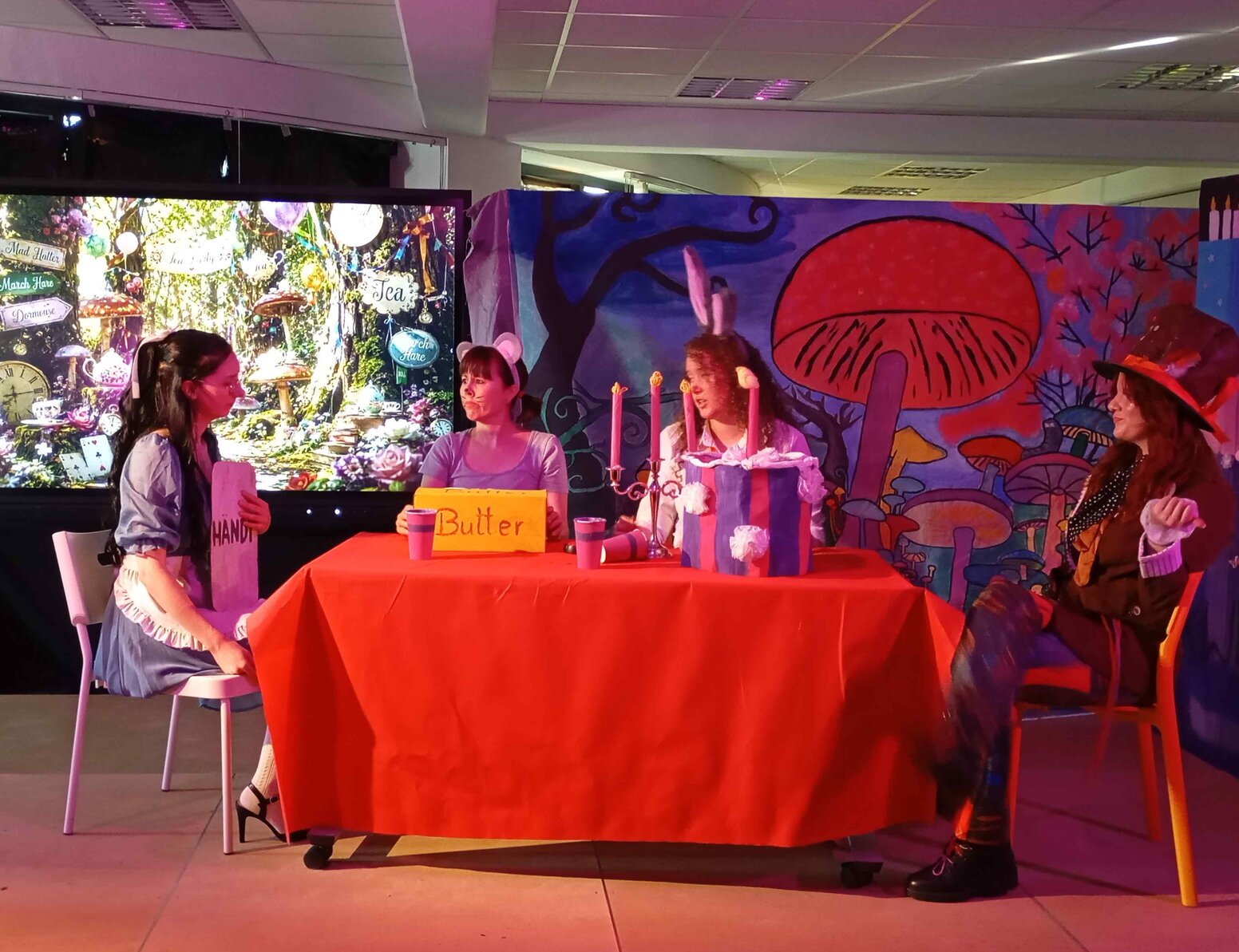 Theaterszene im Stil von „Alice im Wunderland“: Vier kostümierte Personen sitzen an einem rot gedeckten Tisch vor einer fantasievollen Kulisse mit großen Pilzen. Im Hintergrund sind ein Bildschirm mit einem märchenhaften Gartenmotiv sowie ein Schild mit der Aufschrift „Butter“ auf dem Tisch zu sehen.