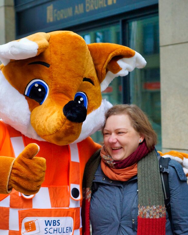 Fuchs-Maskottchen von WBS SCHULEN in karierter Latzhose posiert neben einer Person vor einem Schulgebäude.