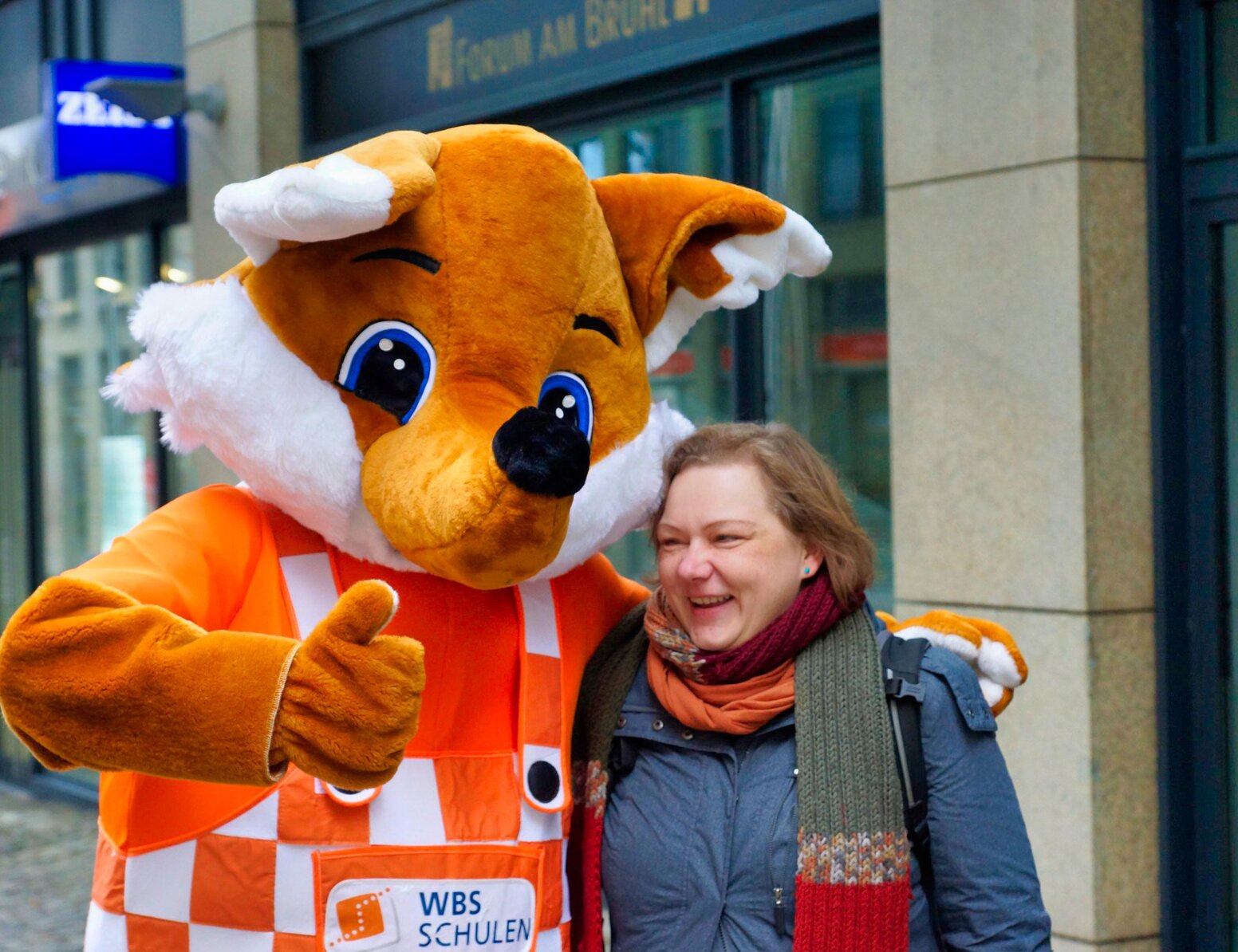 Fuchs-Maskottchen von WBS SCHULEN in karierter Latzhose posiert neben einer Person vor einem Schulgebäude.