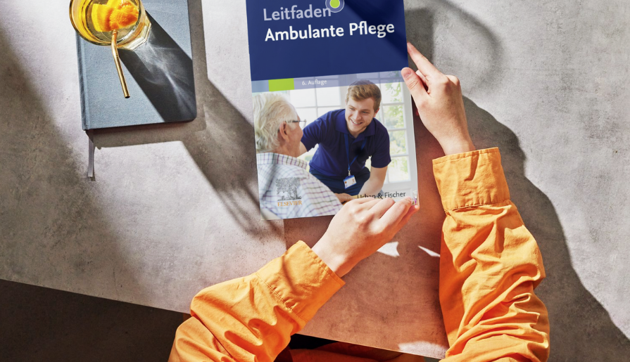 Person mit orangefarbener Kleidung hält den Fachbuch-Titel „Leitfaden Ambulante Pflege, 6. Auflage“ auf einem Tisch. Auf dem Buchcover ist eine Pflegeszene abgebildet; daneben liegen ein Notizbuch und ein Getränk im Sonnenlicht.