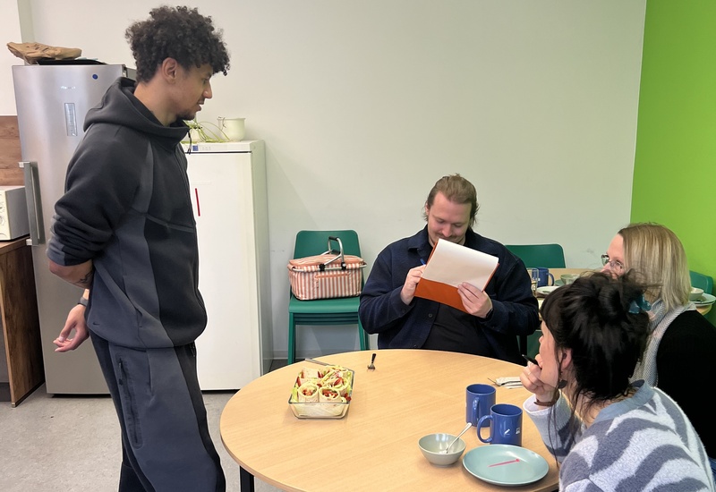 Schüler und Juroren in einer Gemeinschaftsküche mit selbst zubereiteten Snacks auf einem runden Tisch.