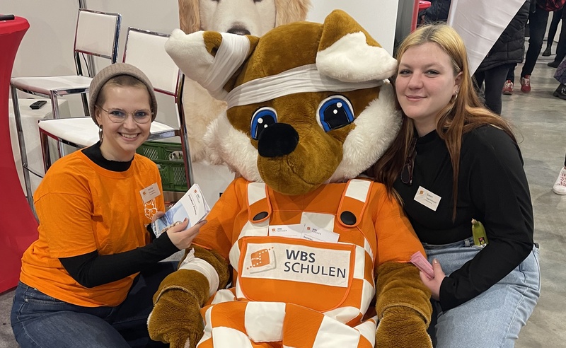 Zwei Auszubildende mit einem Fuchs-Maskottchen am Messestand der WBS SCHULEN bei einer Berufsorientierungsmesse.