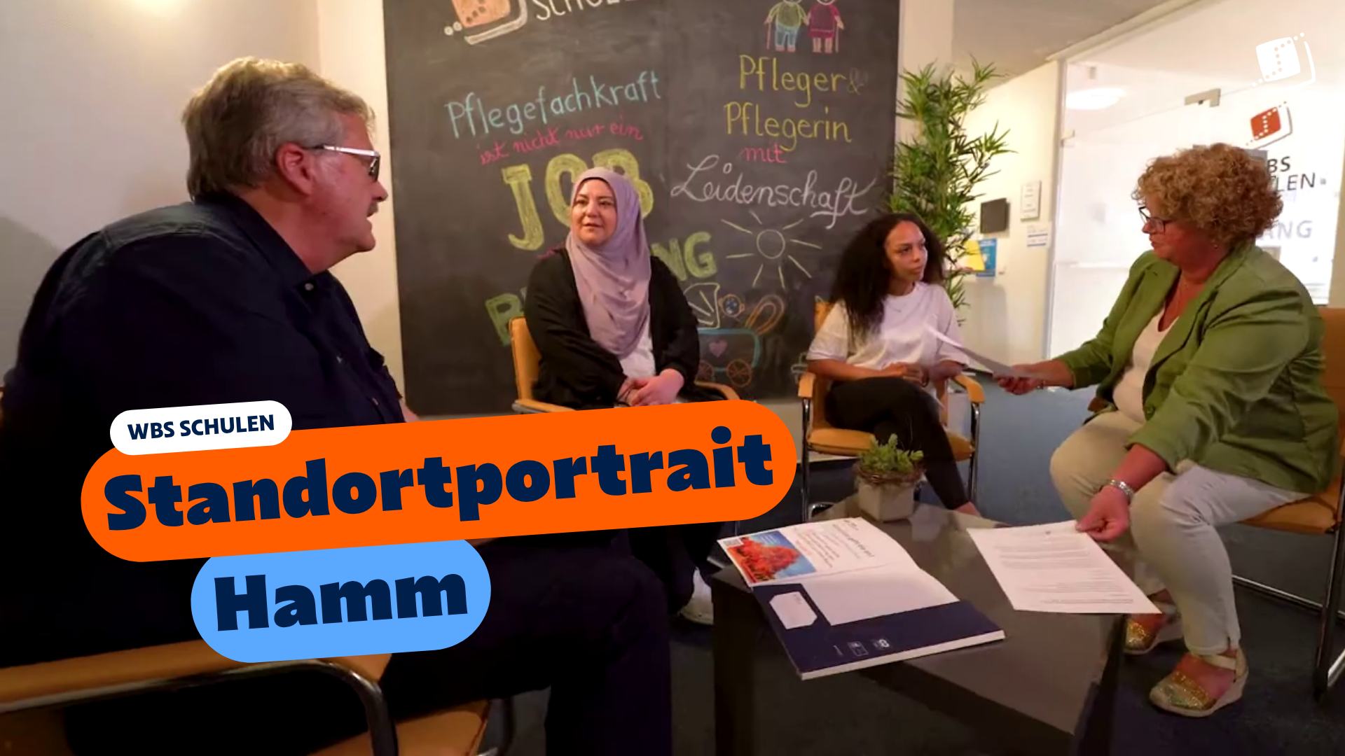 Vier Personen sitzen in einem Besprechungsbereich und sprechen miteinander. Im Vordergrund liegen Unterlagen auf einem Tisch, im Hintergrund ist eine beschriftete Tafel zu sehen. Eingeblendeter Text: „WBS SCHULEN – Standortportrait Hamm“.