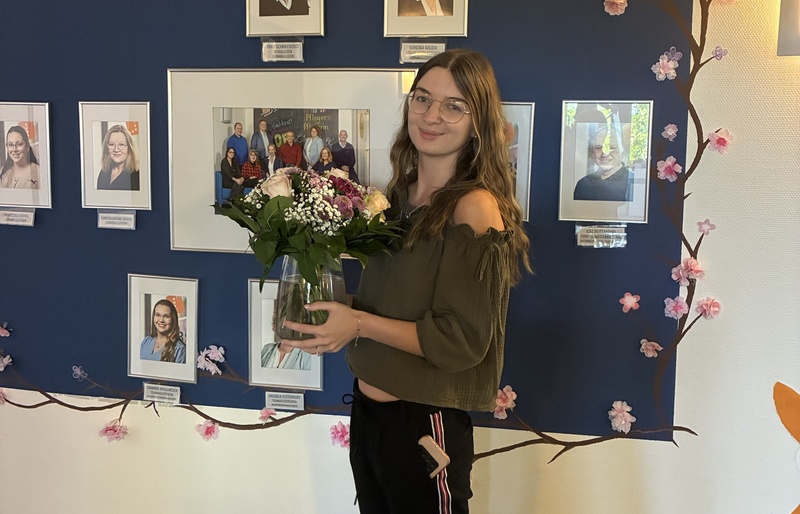 Neue Lernbegleitung mit Blumenstrauß vor einer Fotowand der Mitarbeitenden der WBS SCHULEN.