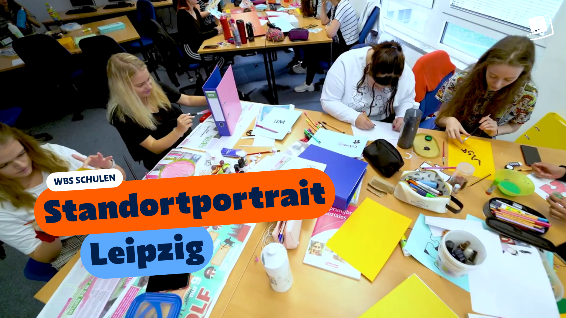 Mehrere Personen sitzen in einem Unterrichtsraum an Gruppentischen und gestalten kreative Projekte mit Papier, Stiften und Bastelmaterialien. Eingeblendeter Text: „WBS SCHULEN – Standortportrait Leipzig“.