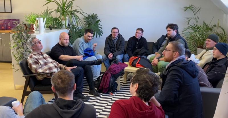 Mehrere Personen sitzen in einem Kreis in einem hellen Aufenthaltsraum und tauschen sich miteinander aus. Die Gruppe ist auf Sofas und Sesseln um einen kleinen Tisch versammelt, umgeben von zahlreichen Zimmerpflanzen.