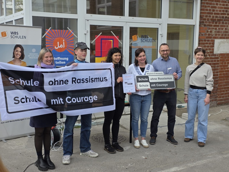 Aufnahmefeier vor einem Schulgebäude: Eine Gruppe präsentiert Banner und Schild „Schule ohne Rassismus – Schule mit Courage“.