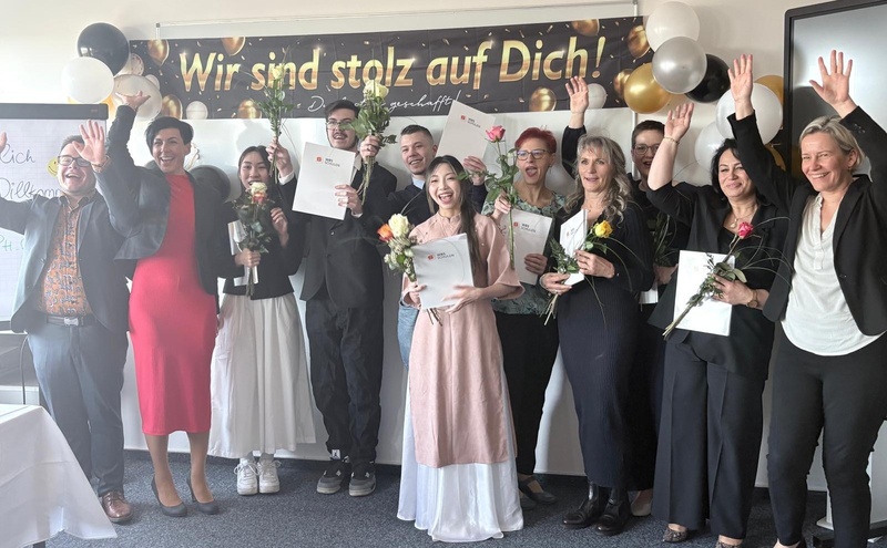 Abschlussfeier einer Pflegehilfe-Ausbildung: Absolventinnen und Absolventen präsentieren ihre Zertifikate und Blumen vor einem Gratulationsbanner.