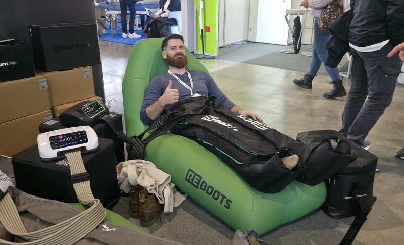 Eine Lehrkraft für Physiotherapie-Ausbildungen testet an einem Messestand ein REBOOTS-Kompressionsmassage-System auf einer grünen Liege.
