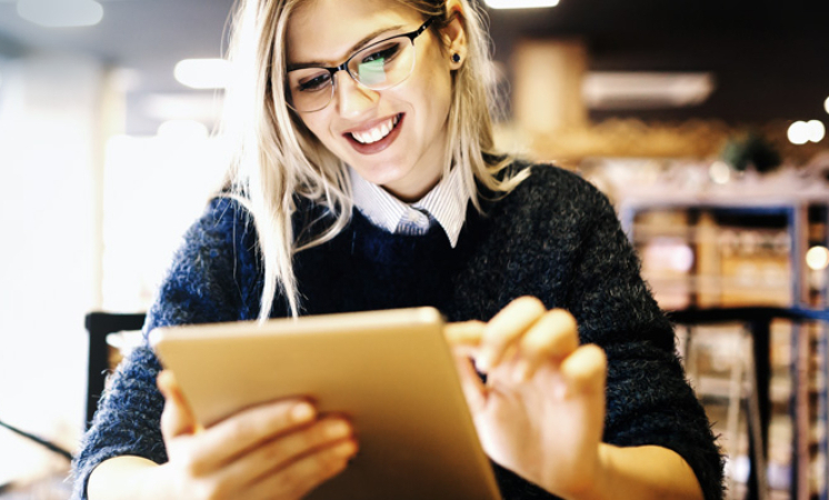 junge, blonde Frau mit Brille schaut sich etwas auf ihrem iPad an.