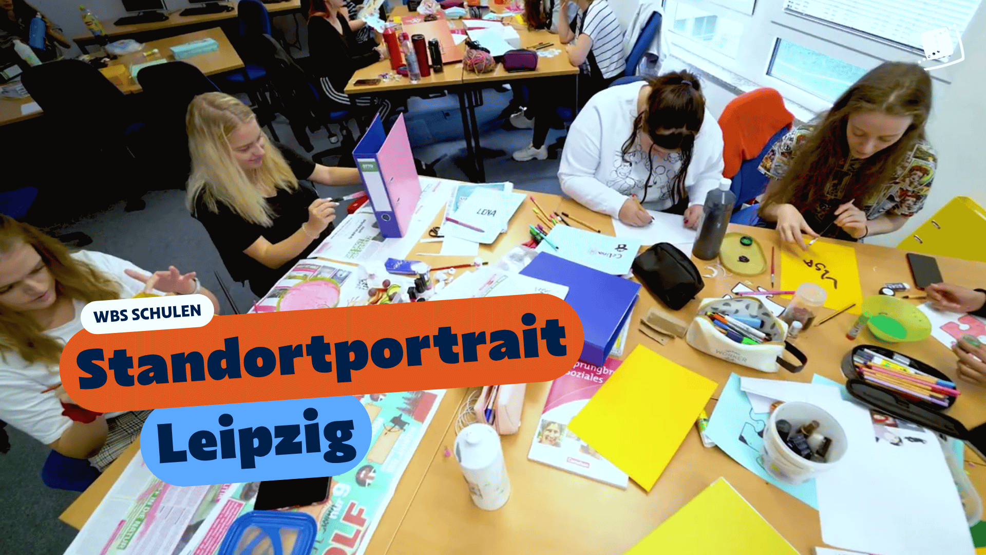 Mehrere Personen sitzen in einem Unterrichtsraum an Gruppentischen und gestalten kreative Projekte mit Papier, Stiften und Bastelmaterialien. Eingeblendeter Text: „WBS SCHULEN – Standortportrait Leipzig“.